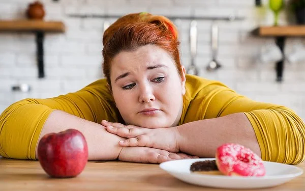 Eine etwas fülligere Frau sitzt mit verschränkten Armen an einem Tisch. Die Frau kann sich nicht zwischen einem Apfel oder einem Donut entscheiden, die vor ihr stehen.