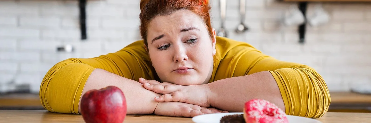 Eine etwas fülligere Frau sitzt mit verschränkten Armen an einem Tisch. Die Frau kann sich nicht zwischen einem Apfel oder einem Donut entscheiden, die vor ihr stehen.