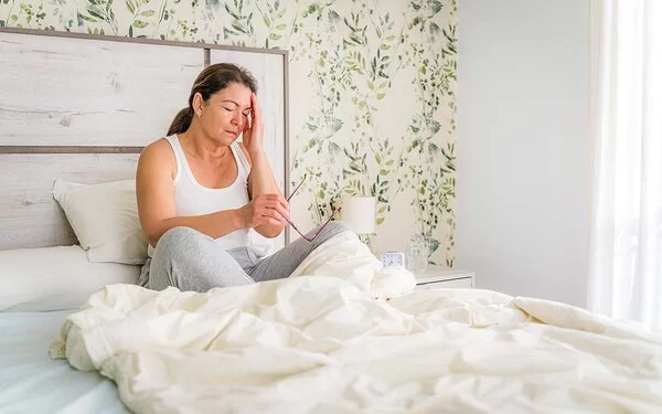 Eine etwas fülligere Frau sitzt mit verschränkten Armen an einem Tisch. Die Frau kann sich nicht zwischen einem Apfel oder einem Donut entscheiden, die vor ihr stehen.