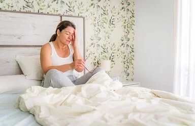 Eine etwas fülligere Frau sitzt mit verschränkten Armen an einem Tisch. Die Frau kann sich nicht zwischen einem Apfel oder einem Donut entscheiden, die vor ihr stehen.