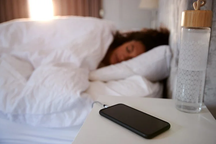 Ein Smartphone wird auf einem Nachttisch aufgeladen. Im Hintergrund schläft eine Frau in ihrem Bett.