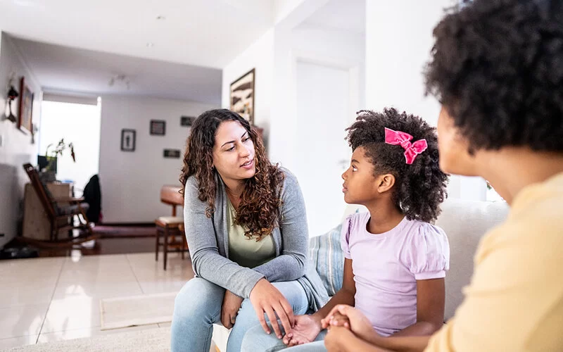 Zwei Frauen sprechen mit ihrer Tochter im Wohnzimmer auf der Couch.