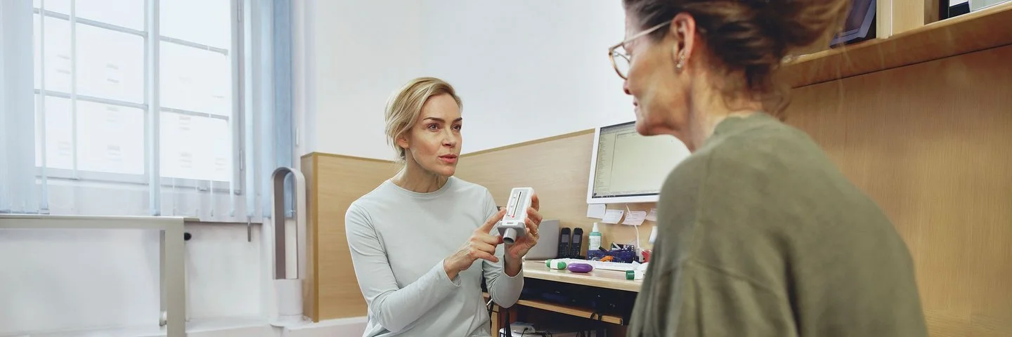 Zwei Frauen sitzen sich in einem hellen Büro gegenüber, eine zeigt auf ein medizinisches Gerät in der Hand.
