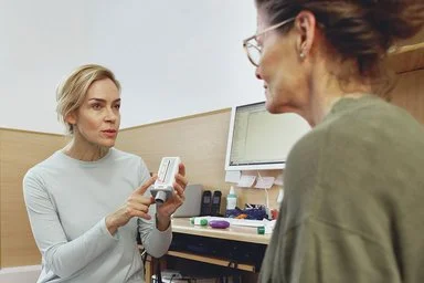 Zwei Frauen sitzen sich in einem hellen Büro gegenüber, eine zeigt auf ein medizinisches Gerät in der Hand.