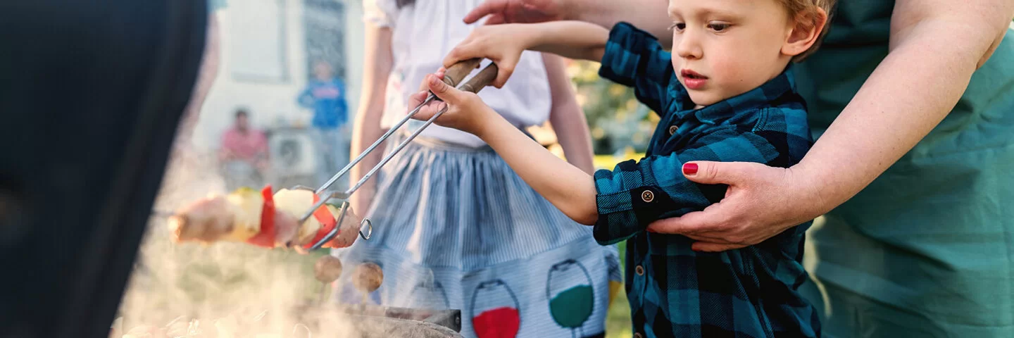 Eine Frau hilft einem kleinen Jungen beim Grillen eines Gemüsespießes. Ein älteres Mädchen schaut zu.