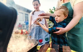 Eine Frau hilft einem kleinen Jungen beim Grillen eines Gemüsespießes. Ein älteres Mädchen schaut zu.