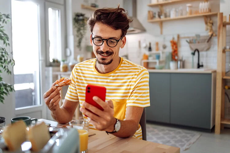 Mann sitzt am Küchentisch beim Frühstück, in der rechten Hand hält er ein Marmeladenbrot, mit der linken ein Smartphone.