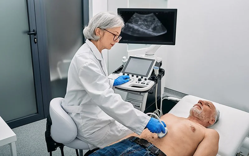 Eine Ärztin führt bei einem älteren, oberkörperfreien Patienten eine Ultraschalluntersuchung des Bauches durch.