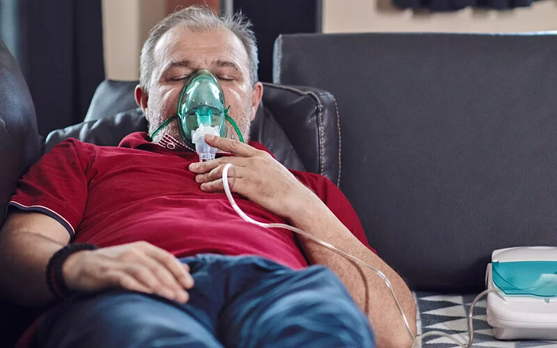 Ein älterer Mann benutzt zuhause auf einem Sofa eine Sauerstoffmaske, die an einen tragbaren Sauerstoffkonzentrator angeschlossen ist.