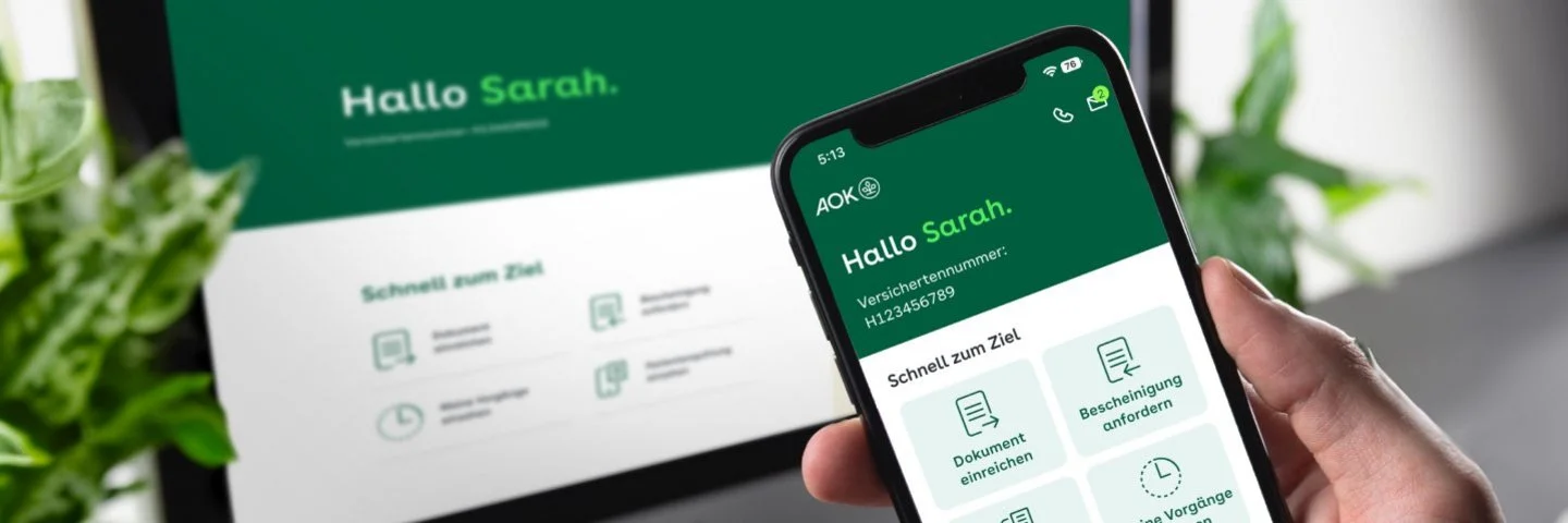 Eine Person hält ein Smartphone in der Hand. Auf dem Bildschirm ist die geöffnete App „Meine AOK“ zu sehen. Im Hintergrund steht ein PC-Monitor, auf dem ebenfalls das Online-Portal „Meine AOK“ zu sehen ist.