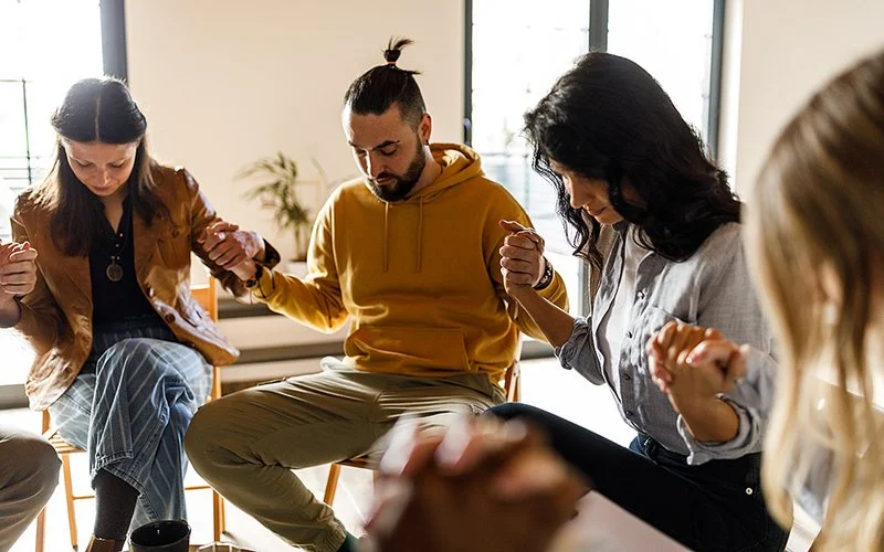 Bei einer Gruppentherapie sitzen die Teilnehmenden in einem Stuhlkreis und halten sich an den Händen. Im Foto ist ein Ausschnitt des Stuhlkreises mit zwei Frauen und zwei Männern abgebildet.