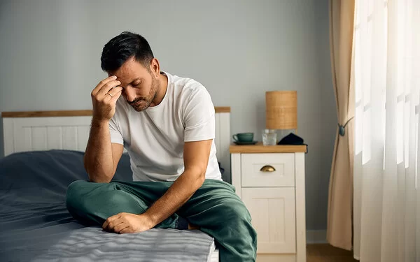 Ein junger Mann sitzt auf seinem Bett im Schlafzimmer. Mit gesenktem Kopf stützt er den rechten Ellenbogen auf dem rechten Knie ab und fasst sich mit der rechten Hand an die Stirn.