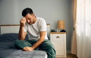 Ein junger Mann sitzt auf seinem Bett im Schlafzimmer. Mit gesenktem Kopf stützt er den rechten Ellenbogen auf dem rechten Knie ab und fasst sich mit der rechten Hand an die Stirn.