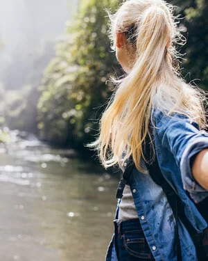 Eine blonde Frau steht im Wald, mit dem Rücken zur Kamera.