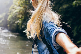 Eine blonde Frau steht im Wald, mit dem Rücken zur Kamera.