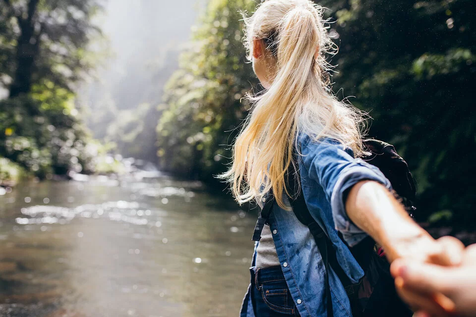 Eine blonde Frau steht im Wald, mit dem Rücken zur Kamera.