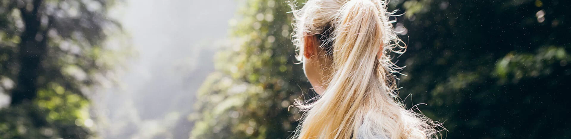 Eine blonde Frau steht im Wald, mit dem Rücken zur Kamera.