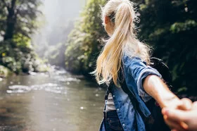 Eine blonde Frau steht im Wald, mit dem Rücken zur Kamera.