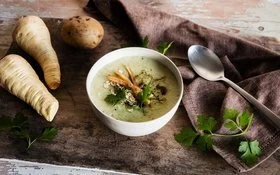 Schale mit cremiger Suppe auf Holztisch, garniert mit Petersilie, Haferflocken und gerösteten Pastinaken; daneben Pastinaken, eine Kartoffel, Löffel und Tuch.