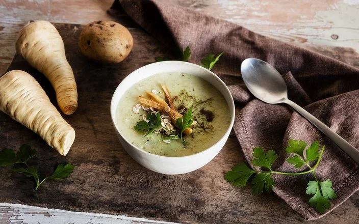 Schale mit cremiger Suppe auf Holztisch, garniert mit Petersilie, Haferflocken und gerösteten Pastinaken; daneben Pastinaken, eine Kartoffel, Löffel und Tuch.
