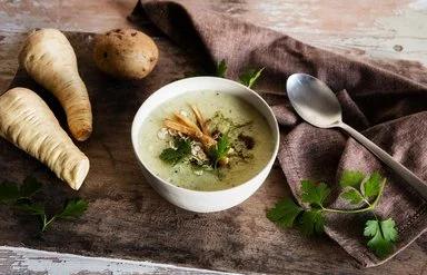 Schale mit cremiger Suppe auf Holztisch, garniert mit Petersilie, Haferflocken und gerösteten Pastinaken; daneben Pastinaken, eine Kartoffel, Löffel und Tuch.
