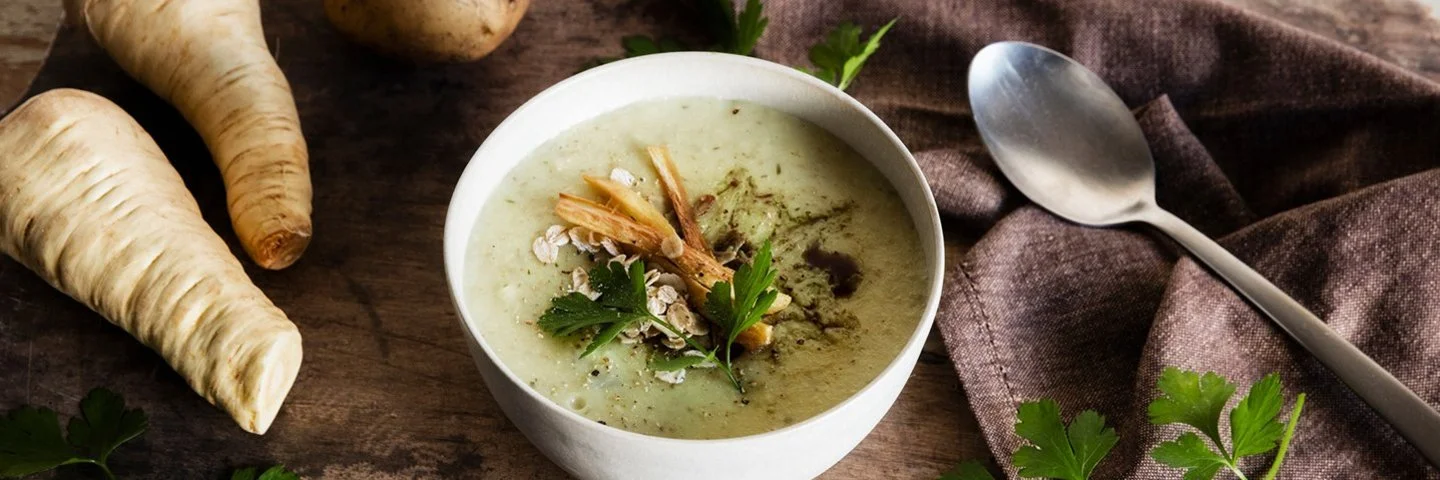Schale mit cremiger Suppe auf Holztisch, garniert mit Petersilie, Haferflocken und gerösteten Pastinaken; daneben Pastinaken, eine Kartoffel, Löffel und Tuch.