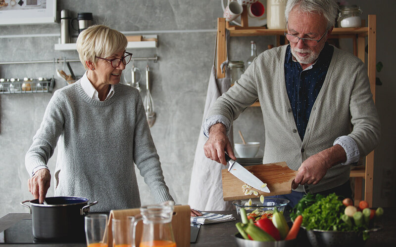 Ein Seniorenpaar steht gemeinsam in der Küche und kocht. Während der Senior Gemüse zurechtschneidet, schaut die Seniorin zu ihm rüber und rührt mit einem Kochlöffel im Topf herum.