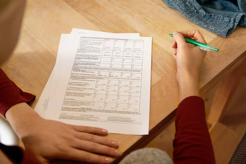 Eine Frau sitzt an einem Tisch mit mehreren Zetteln vor sich. In der Hand hält sie einen Stift. Die AOK stellt alle wichtigen Leistungsanträge und Formulare zusammen.