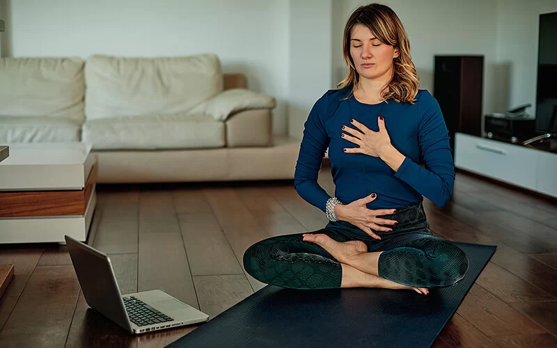 Eine Frau sitzt auf einer Yogamatte auf dem Boden und macht eine Atemübung. Mit geschlossenen Augen atmet sie tief ein und spürt dabei in den Bauch hinein.