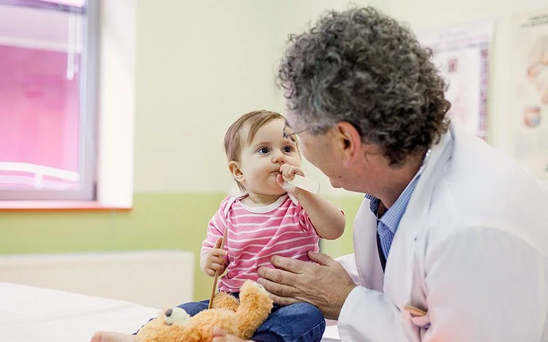 In einem Sprechzimmer sitzt ein Kleinkind mit einem Teddy und führt einen Spatel an den Mund. Ein Mann in weißem Kittel sitzt daneben und schaut das Kind an.