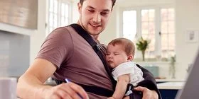Ein junger Mann sitzt zu Hause an seinem Schreibtisch und füllt ein Formular aus. Vor seiner Brust hält er sein Baby in einer Trage.