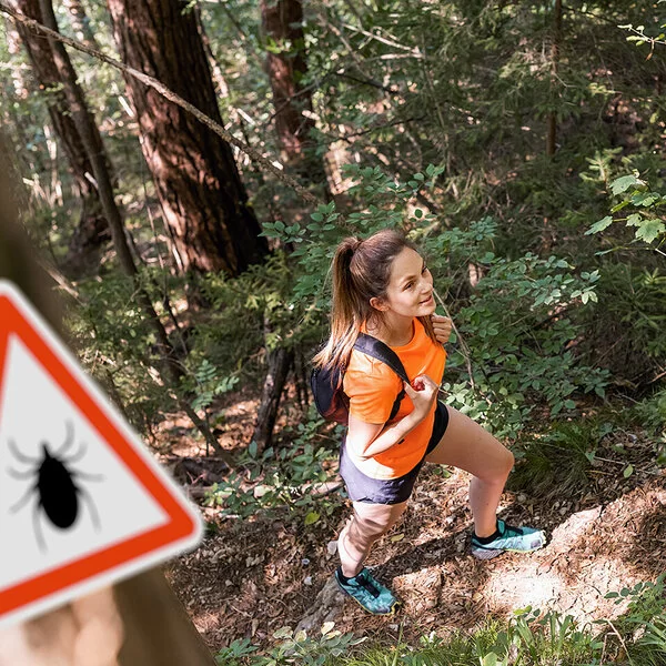Eine junge Frau in robuster Wanderkleidung mit Rücksäcken geht gut gelaunt durch einen dichten Wald.