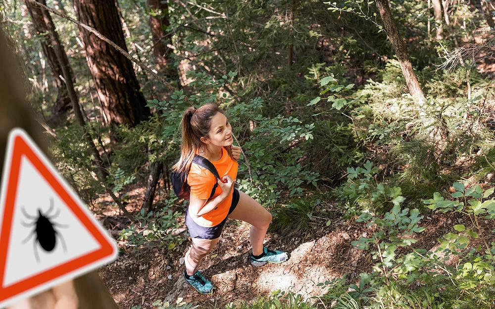 Eine junge Frau in robuster Wanderkleidung mit Rücksäcken geht gut gelaunt durch einen dichten Wald.