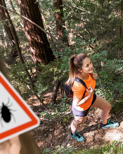 Eine junge Frau in robuster Wanderkleidung mit Rücksäcken geht gut gelaunt durch einen dichten Wald.