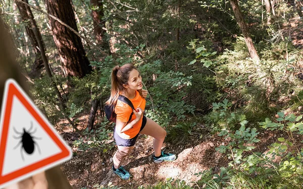 Eine junge Frau in robuster Wanderkleidung mit Rücksäcken geht gut gelaunt durch einen dichten Wald.