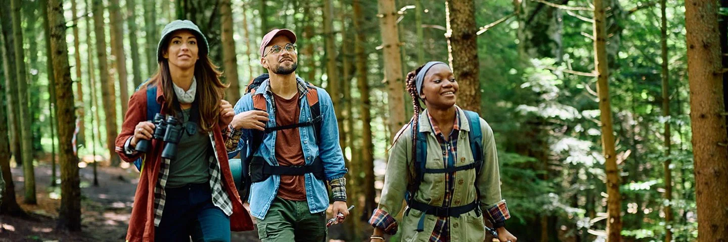 Drei junge Leute in robuster Wanderkleidung mit Rücksäcken gehen gut gelaunt durch einen dichten Wald.