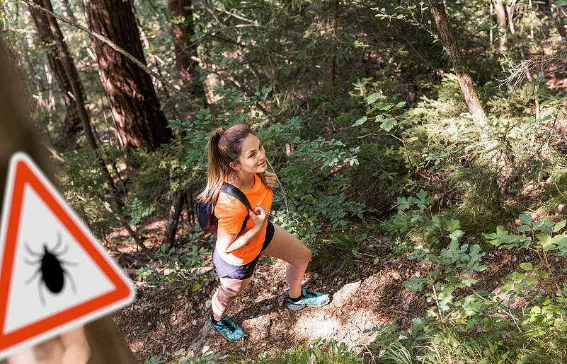 Eine junge Frau in robuster Wanderkleidung mit Rücksäcken geht gut gelaunt durch einen dichten Wald.