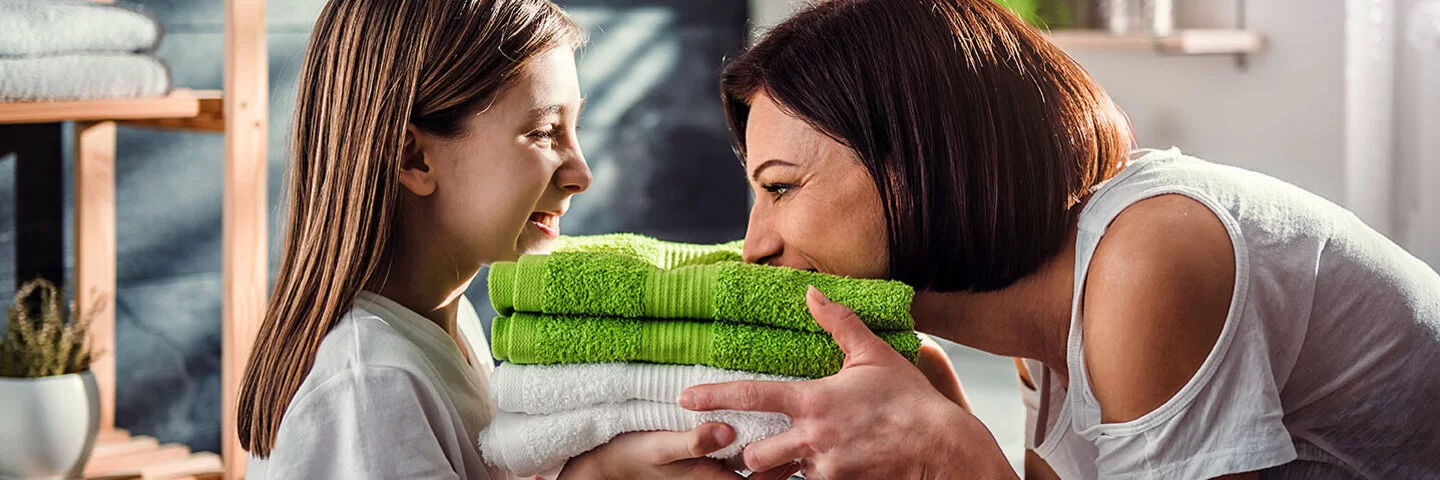 Eine Mutter steht mit ihrer Tochter vor der Waschmaschine im Bad. Sie halten frisch gewaschene, zusammengelegte Handtücher in den Händen, die Mutter riecht daran, beide lachen.