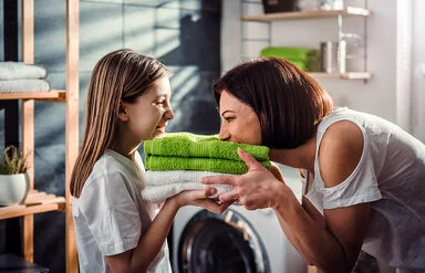 Eine Mutter steht mit ihrer Tochter vor der Waschmaschine im Bad. Sie halten frisch gewaschene, zusammengelegte Handtücher in den Händen, die Mutter riecht daran, beide lachen.