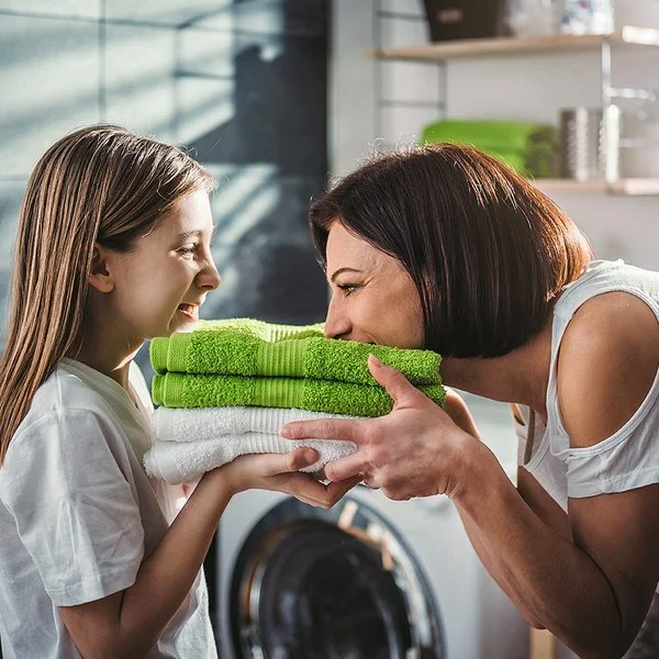 Eine Mutter steht mit ihrer Tochter vor der Waschmaschine im Bad. Sie halten frisch gewaschene, zusammengelegte Handtücher in den Händen, die Mutter riecht daran, beide lachen.
