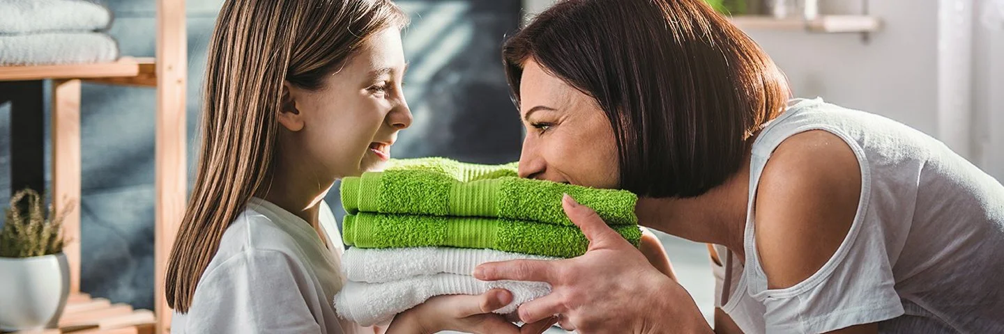 Eine Mutter steht mit ihrer Tochter vor der Waschmaschine im Bad. Sie halten frisch gewaschene, zusammengelegte Handtücher in den Händen, die Mutter riecht daran, beide lachen.