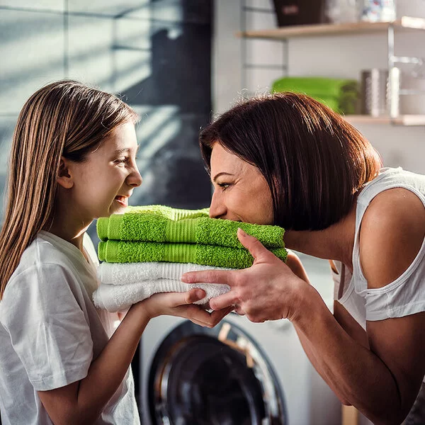 Eine Mutter steht mit ihrer Tochter vor der Waschmaschine im Bad. Sie halten frisch gewaschene, zusammengelegte Handtücher in den Händen, die Mutter riecht daran, beide lachen.