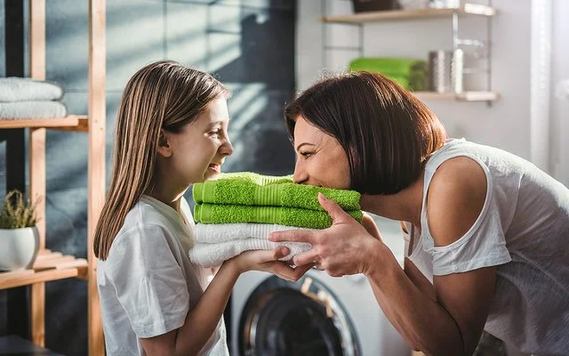 Eine Mutter steht mit ihrer Tochter vor der Waschmaschine im Bad. Sie halten frisch gewaschene, zusammengelegte Handtücher in den Händen, die Mutter riecht daran, beide lachen.