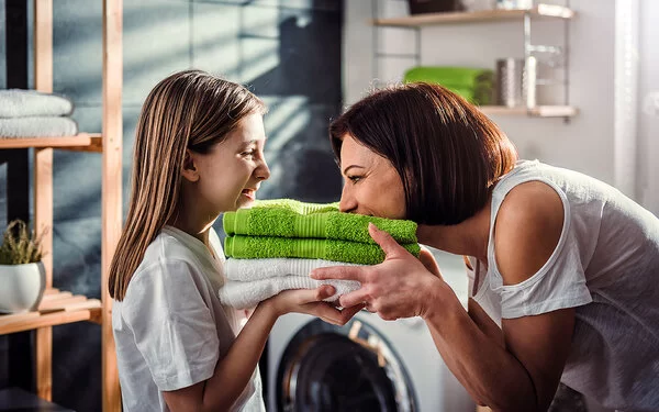 Eine Mutter steht mit ihrer Tochter vor der Waschmaschine im Bad. Sie halten frisch gewaschene, zusammengelegte Handtücher in den Händen, die Mutter riecht daran, beide lachen.