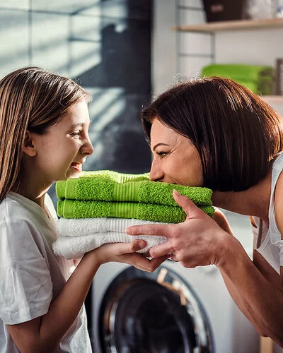 Eine Mutter steht mit ihrer Tochter vor der Waschmaschine im Bad. Sie halten frisch gewaschene, zusammengelegte Handtücher in den Händen, die Mutter riecht daran, beide lachen.