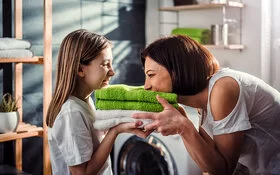 Eine Mutter steht mit ihrer Tochter vor der Waschmaschine im Bad. Sie halten frisch gewaschene, zusammengelegte Handtücher in den Händen, die Mutter riecht daran, beide lachen.
