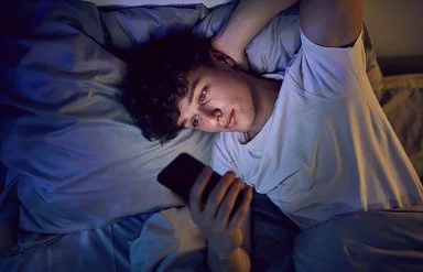 Ein Jugendlicher liegt im Bett und schaut auf sein Handy.