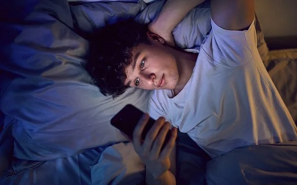 Ein Jugendlicher liegt im Bett und schaut auf sein Handy.