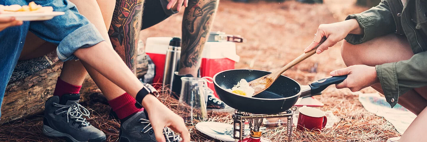 Eine Gruppe junger Menschen sitzt um einen Campinggrill und probiert Camping Rezepte aus.