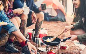 Eine Gruppe junger Menschen sitzt um einen Campinggrill und probiert Camping Rezepte aus.